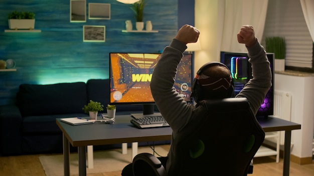 Jugador de videojuego levantando las manos después de ganar la competencia de disparos en primera persona usando auriculares. Jugador profesional profesional que juega videojuegos en línea con nuevos gráficos en una computadora potente
