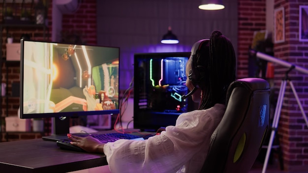 Una jugadora afroamericana que usa la configuración de juegos de PC para pasar un buen rato jugando un juego de acción en línea multijugador en la sala de estar de su casa. Mujer transmitiendo disparos en primera persona mientras habla con auriculares.