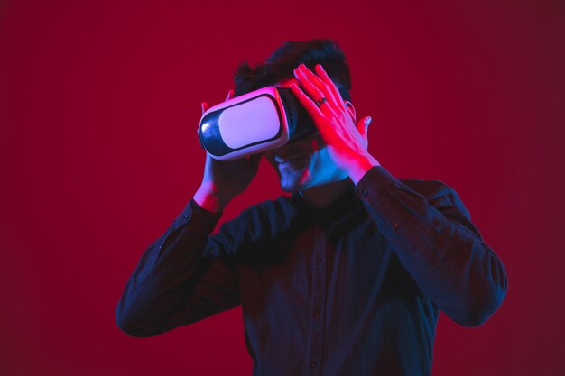 Jugando usaba cascos de realidad virtual. Retrato de joven caucásico aislado en la pared roja en luz de neón. Hermosa modelo. Concepto de emociones humanas, expresión facial, juventud, dispositivos.