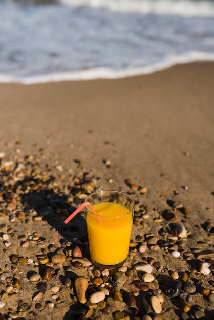 Jugo amarillo en vaso con pajita roja cerca de la orilla del mar en la playa