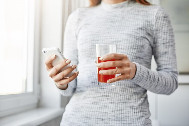 El jugo de la mañana refresca la mente. Retrato recortado de mujer delgada moderna de pie junto a la ventana, sosteniendo un vaso de jugo y navegando en la red a través de un teléfono inteligente. Chica quiere llamar a su jefe y tomarse el día libre