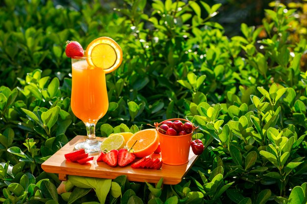 Jugo de naranja en una copa con cítricos, fresa, cereza, vista lateral de la tabla de cortar en un prado