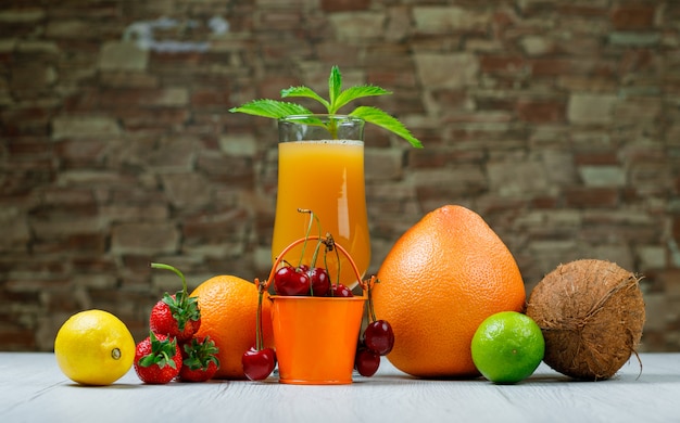 Jugo de naranja con menta, naranja, lima, limón, fresa, cereza, coco, pomelo en una copa sobre piedra de ladrillo y superficie de madera, vista lateral.