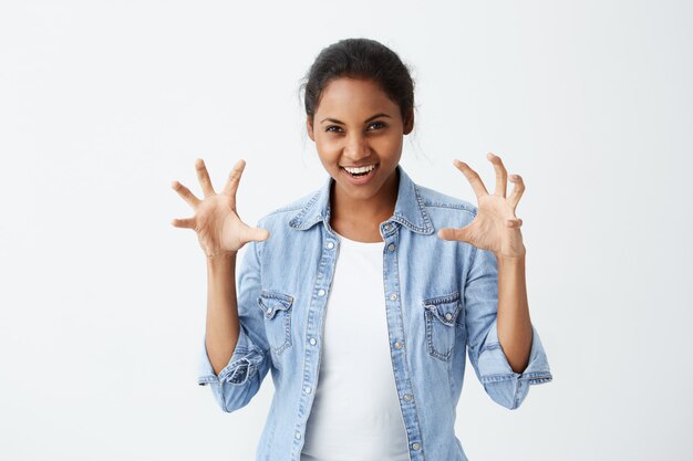 Juguetón divirtiéndose Mujer afroamericana con cabello oscuro vestido informalmente, mostrando sus dientes gesticulando activamente, tratando de asustar a alguien. Lenguaje corporal.