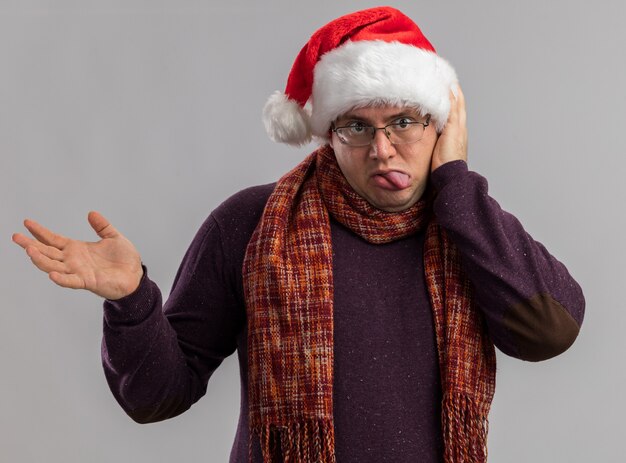 Juguetón hombre adulto con gafas y gorro de Papá Noel con bufanda alrededor del cuello manteniendo la mano en la cabeza mostrando la lengua y la mano vacía aislada en la pared blanca
