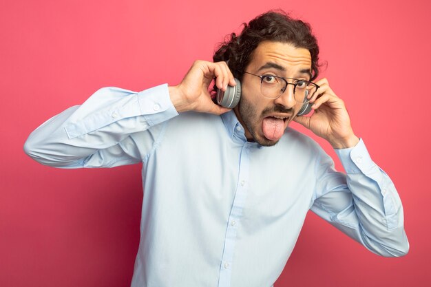 Juguetón joven apuesto hombre caucásico con gafas y auriculares agarrando auriculares mostrando la lengua mirando a cámara aislada sobre fondo carmesí