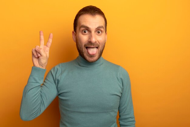 Juguetón joven caucásico mostrando la lengua haciendo el signo de la paz aislado en la pared naranja con espacio de copia