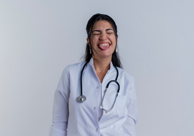 Juguetón joven doctora vistiendo bata médica y un estetoscopio mostrando la lengua con los ojos cerrados aislados