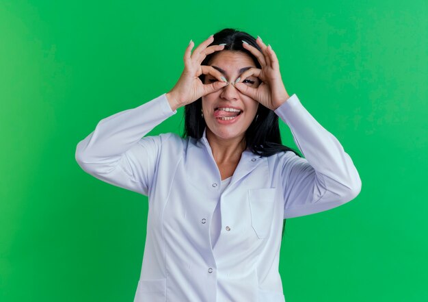 Juguetón joven doctora vistiendo bata médica mirando mostrando la lengua haciendo gesto de mirada con las manos como binoculares