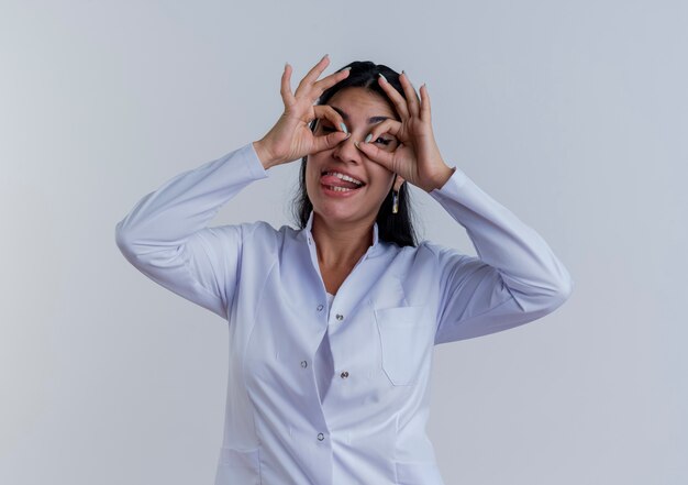 Juguetón joven doctora vistiendo bata médica mostrando la lengua haciendo gesto de mirada mirando aislado