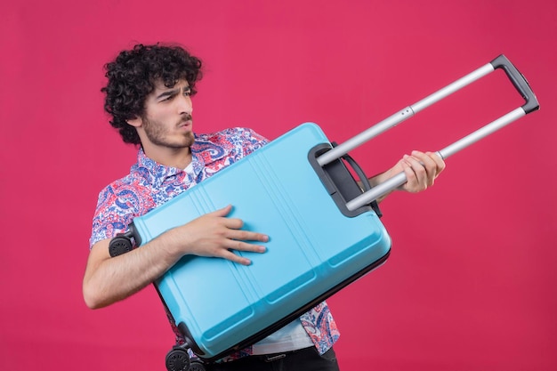 Foto gratuita juguetón joven guapo viajero rizado hombre sosteniendo la maleta y usándola como guitarra en el espacio rosa aislado