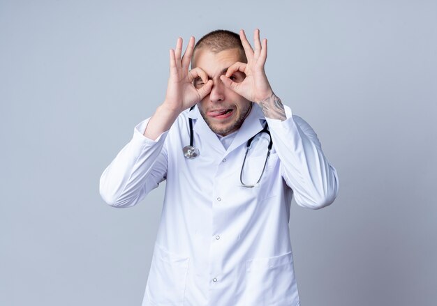 Juguetón joven médico vistiendo una bata médica y un estetoscopio alrededor de su cuello haciendo gesto de mirada guiñando un ojo y mostrando la lengua aislada sobre fondo blanco con espacio de copia