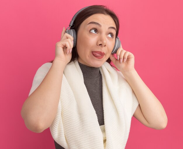 Juguetón joven mujer bonita vistiendo y agarrando auriculares mostrando la lengua mirando al lado aislado en la pared rosa