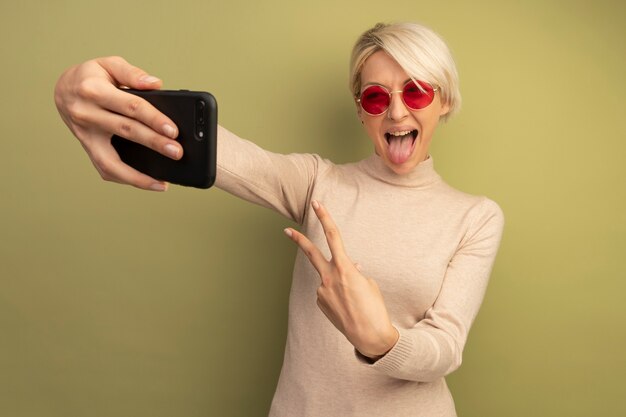 Juguetón joven rubia con gafas de sol haciendo el signo de la paz mostrando la lengua tomando selfie aislado en la pared verde oliva