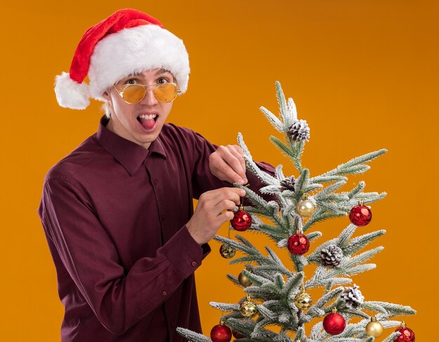 Juguetón joven rubio con gorro de Papá Noel y gafas de pie en la vista de perfil cerca del árbol de Navidad que lo decora con adornos navideños mirando a la cámara mostrando la lengua aislada sobre fondo naranja