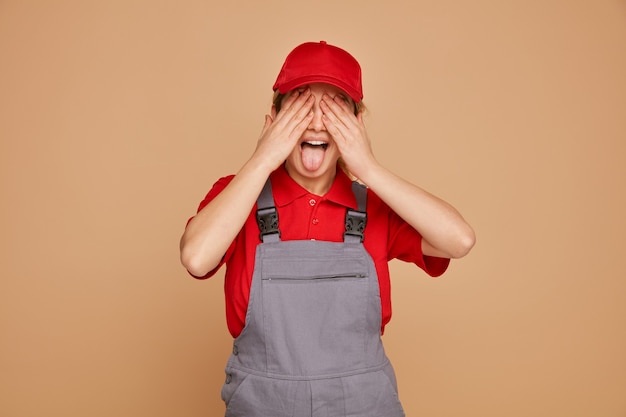 Juguetón joven trabajador de la construcción con uniforme y gorra que cubre los ojos con las manos mostrando la lengua