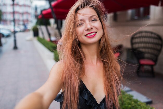 Juguetón lindo rojo escucha a la mujer sonriendo haciendo autorretrato y disfrutando de las vacaciones de verano en Europa. Imagen exterior positiva. Vestido negro, labios rojos.