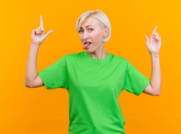 Juguetón mujer eslava rubia de mediana edad mirando a la cámara mostrando la lengua apuntando hacia arriba aislado sobre fondo amarillo