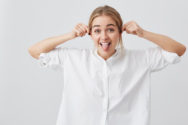 Juguetona chica bonita divertida con expresión sombría lengua demoníaca, tirando de las orejas, burlándose. Juguetón hermosa mujer europea con pelo rubio posando.