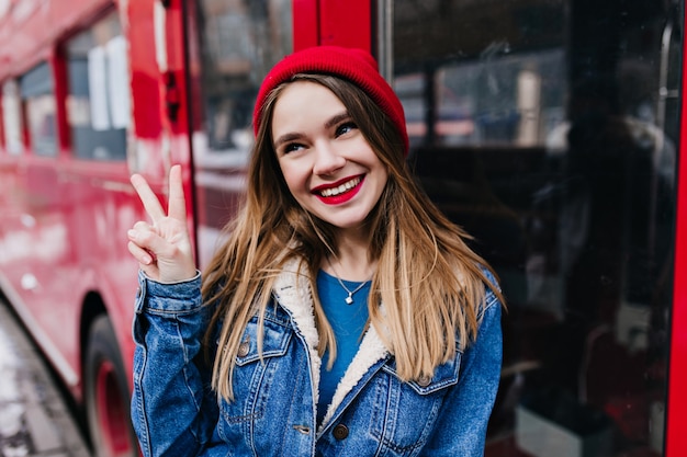 Juguetona chica caucásica con peinado de moda posando con el signo de la paz. Tiro al aire libre de una mujer bonita en chaqueta de mezclilla riendo durante la sesión de fotos.