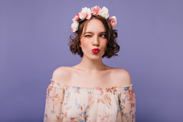 Juguetona chica morena en diadema de flores de pie. Filmación en interiores de mujer jocund con cabello ondulado posando en lindo vestido.