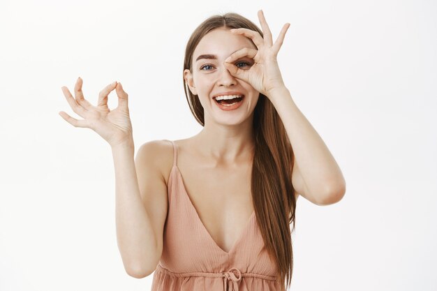 Juguetona y gentil mujer europea en un vestido beige de moda haciendo un buen gesto sobre el ojo y orinando a través del agujero en los dedos sonriendo alegremente divirtiéndose y pasando tiempo divertido sobre una pared gris