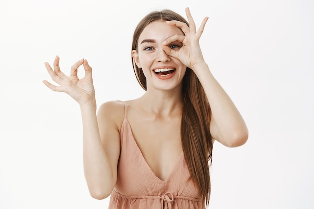 Juguetona y gentil mujer europea en un vestido beige de moda haciendo un buen gesto sobre el ojo y orinando a través del agujero en los dedos sonriendo alegremente divirtiéndose y pasando tiempo divertido sobre una pared gris