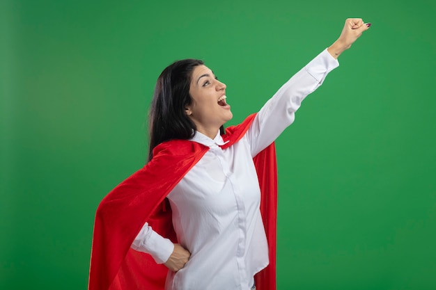 Juguetona joven superhéroe caucásica de pie en la vista de perfil en la pose de superman levantando el puño y mirando a la esquina manteniendo la mano en la cintura aislada sobre fondo verde con copia espacio