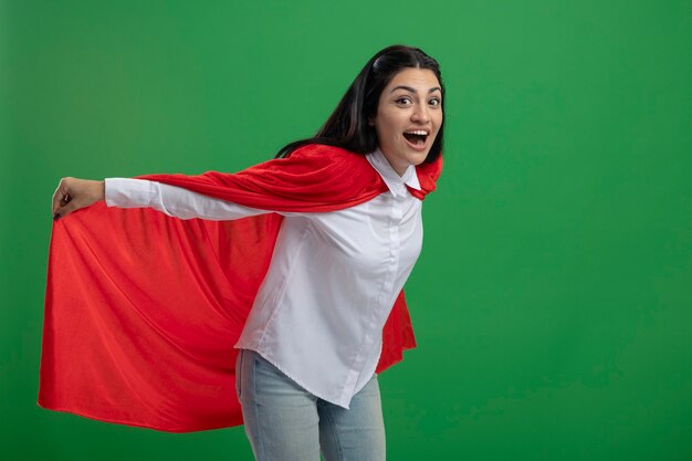 Juguetona joven superhéroe caucásica sosteniendo su capa de héroe y representando vuelo mirando a cámara aislada sobre fondo verde con espacio de copia