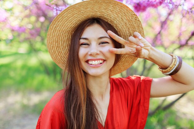 Juguetona linda chica haciendo muecas y mostrando signos, posando en el parque de primavera en árboles florecientes