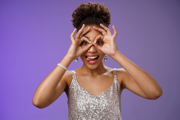 Juguetona mujer africana guapa divertida en peinado afro con vestido plateado brillante divirtiéndose mostrar lengua sonriendo ampliamente hacer bien gesto ok en los ojos se ven emocionados, de pie fondo azul.