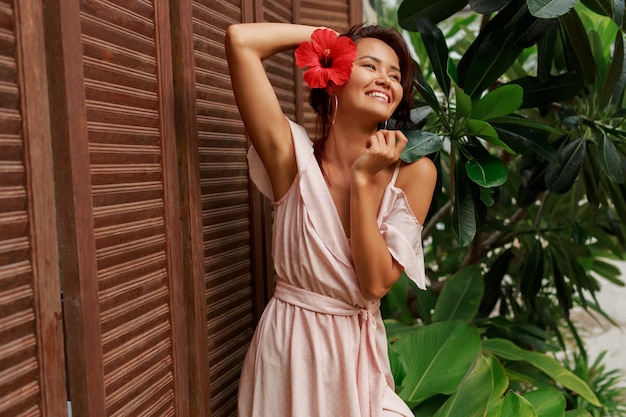 Juguetona mujer asiática con flor de hibisco en pelos y vestido rosa posando en el lujoso resort tropical.