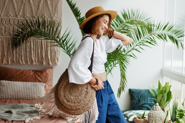 Juguetona mujer bohemia posando en un elegante dormitorio con interiores increíbles, palmeras y macramé