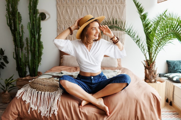 Foto gratuita juguetona mujer bohemia posando en un elegante dormitorio con interiores increíbles, palmeras y macramé