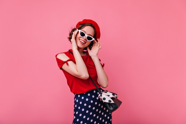 Juguetona mujer francesa posando con gafas de sol. Atractiva chica de pelo oscuro con boina roja sonriendo en la pared rosada.