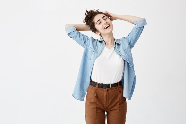 Juguetona mujer joven con cabello oscuro en moño con camisa vaquera con mangas enrolladas riéndose de una buena broma, su mirada y expresión llenas de alegría y felicidad. Concepto de personas y estilo de vida