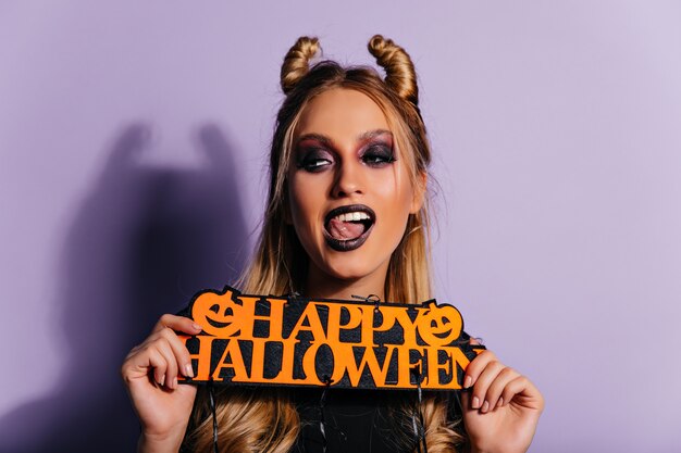 Juguetona mujer joven caucásica disfrutando de la sesión de fotos de halloween. Chica rubia en traje de vampiro posando con decoración de fiesta.