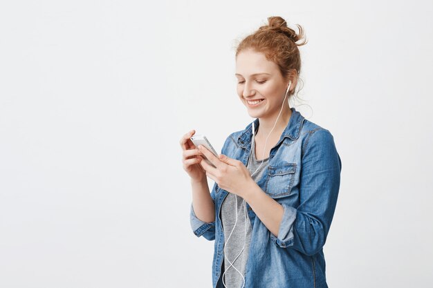 Juguetona mujer pelirroja atractiva con moño, riendo mientras mira la pantalla del teléfono inteligente, mensajes de texto y escuchando música en los auriculares