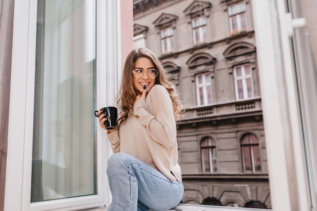 Juguetona mujer de pelo castaño sentada junto a una ventana grande con una sonrisa inspirada