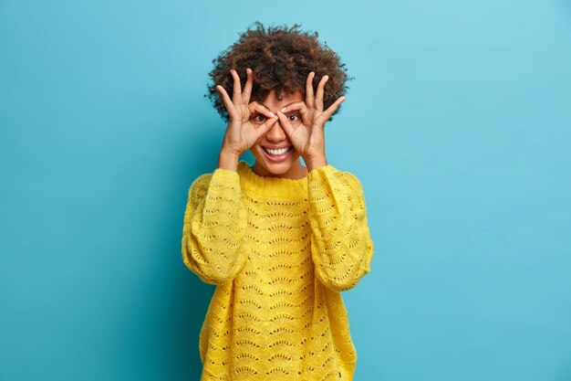 Juguetona mujer de pelo rizado se divierte y hace gafas con los dedos sonríe ampliamente tiene dientes blancos viste un suéter amarillo siendo infantil u optimista vestida con un suéter amarillo se encuentra contra la pared azul