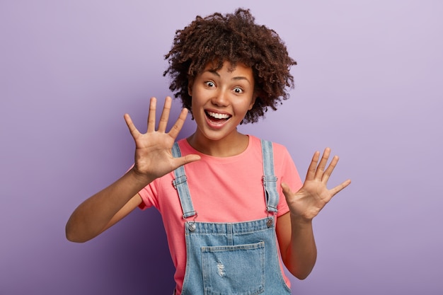 Juguetona mujer de piel oscura divertida muestra las palmas en la cámara, tiene peinado afro, tiene expresión facial alegre