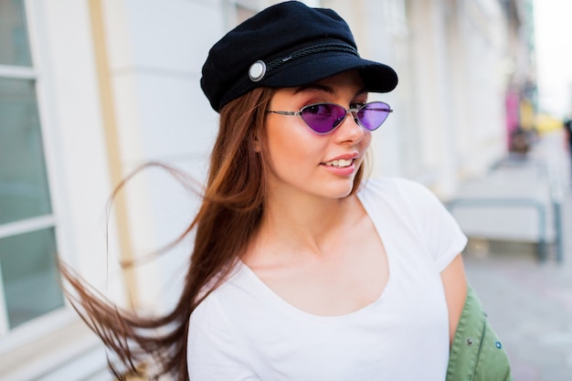 Juguetona mujer posando al aire libre. Lleva gafas retro moradas de moda, gorra negra