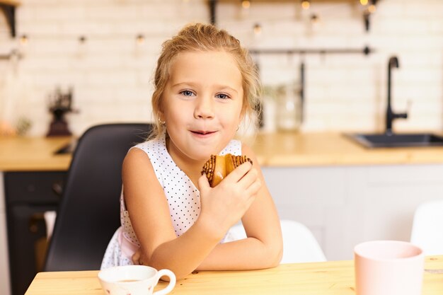 Juguetona niña feliz sentada en la acogedora cocina comiendo sabrosas galletas con tazas en la mesa de comedor. Lindo bebé caucásico divertido masticar pastel dulce horneado con placer y disfrute
