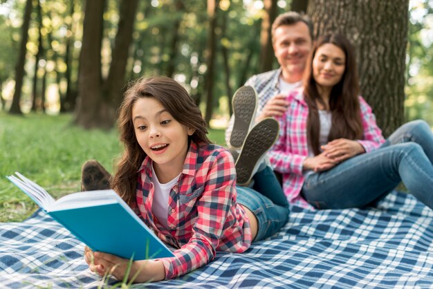 Junte sentarse detrás de su muchacha linda que miente en el libro de lectura de la manta en parque