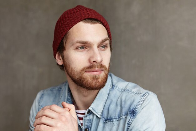 Juventud y felicidad. Cierre plano de atractivo joven europeo con barba con sombrero de moda y camisa de mezclilla mirando a otro lado con alegre expresión facial, pensando en algo agradable