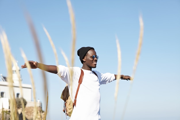 Juventud, felicidad, libertad y viajar. Alegre joven viajero afroamericano con sombrero y gafas de sol extendiendo los brazos y sonriendo felizmente, sintiéndose relajado y en armonía con la naturaleza