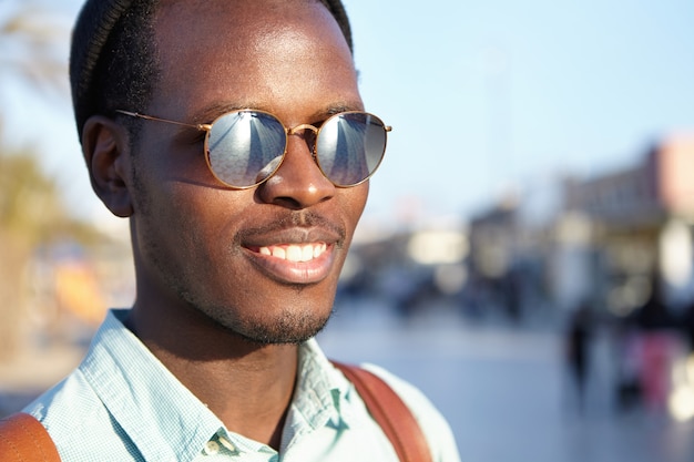 Juventud y felicidad. Personas y estilo de vida. Cierre al aire libre, foto muy detallada de un atractivo joven africano con la espalda sonriendo felizmente, disfrutando de un buen día y un clima agradable, caminando por las calles de la ciudad