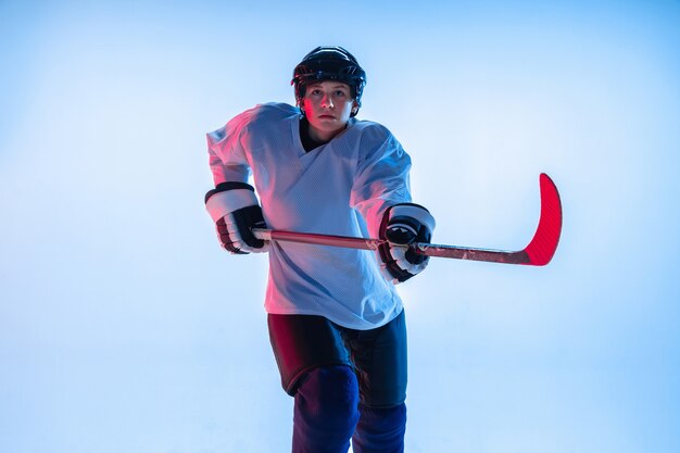 Juventud. Jugador de hockey masculino joven con el palo en la pared blanca con luz de neón. Deportista con equipo y casco practicando. Concepto de deporte, estilo de vida saludable, movimiento, movimiento, acción.