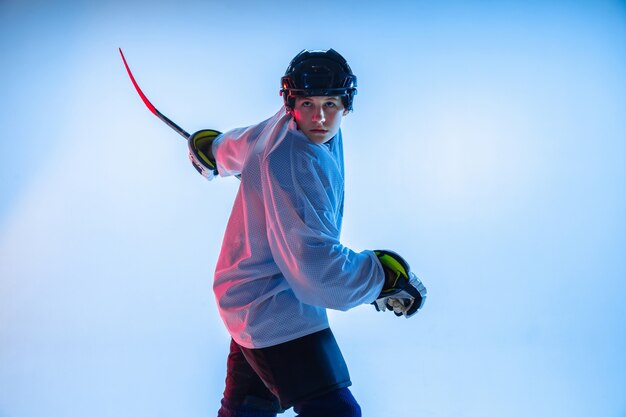 Juventud. Jugador de hockey masculino joven con el palo en la pared blanca con luz de neón. Deportista con equipo y casco practicando. Concepto de deporte, estilo de vida saludable, movimiento, movimiento, acción.