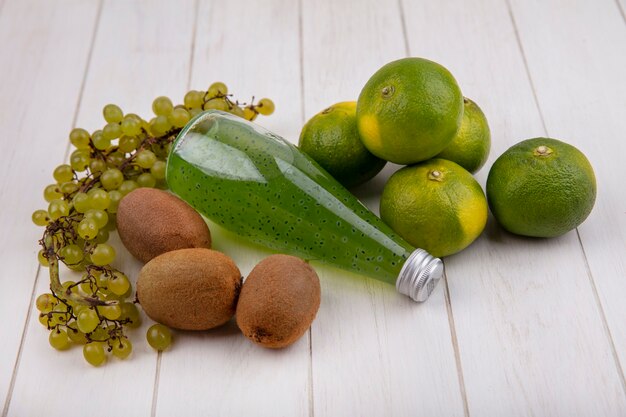 Kiwi de vista lateral con una botella de jugo de uvas y mandarinas en una pared blanca